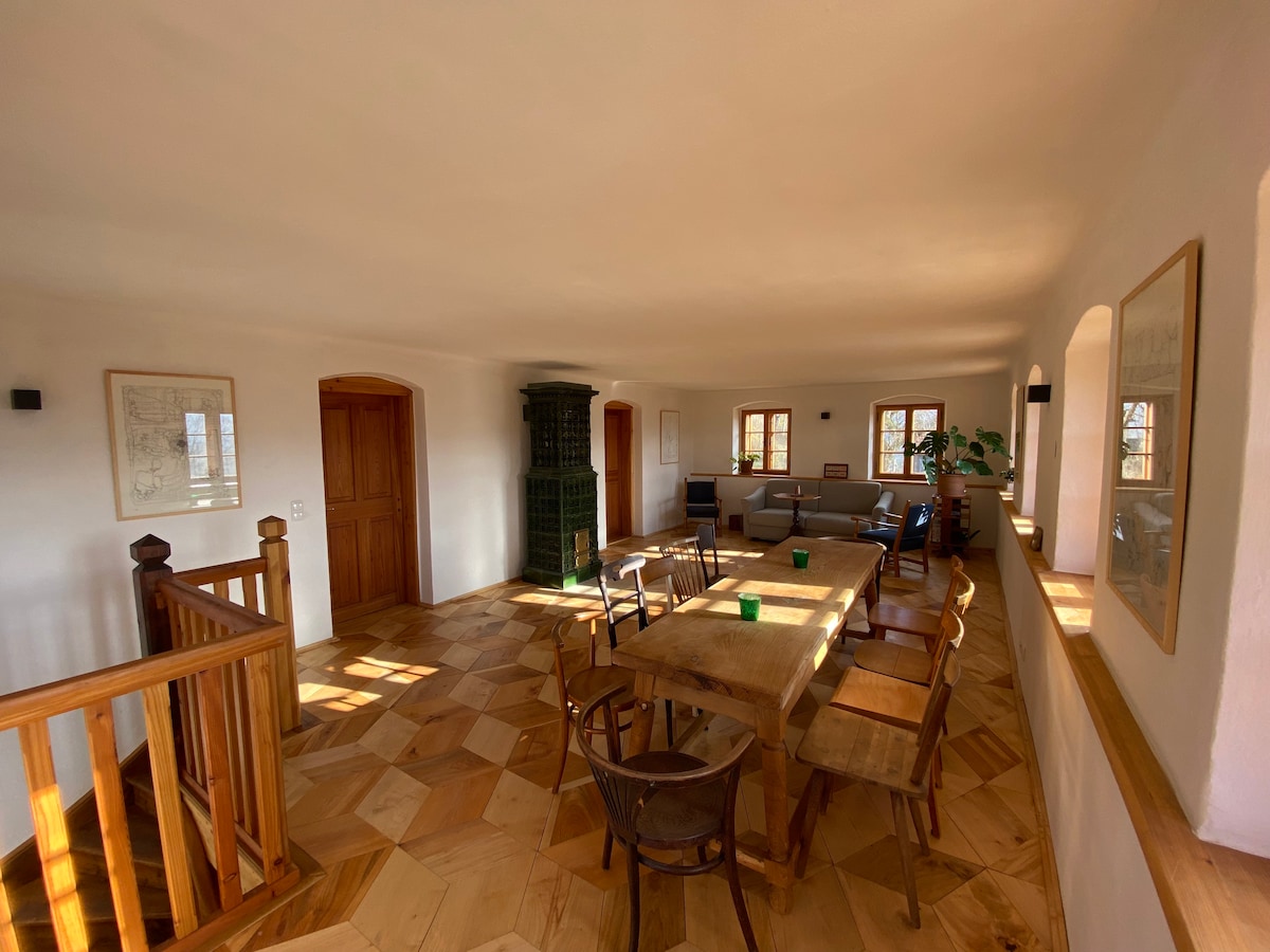 A spacious common area features a large wooden dining table surrounded by several chairs. Natural light enters through multiple windows, illuminating the warm wooden floors. A green ceramic stove stands in the background, complementing the room's neutral tones and offering a cozy touch.