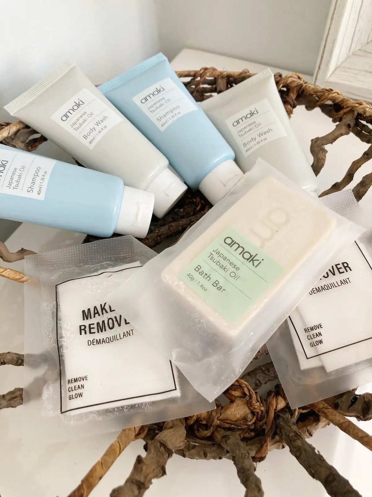An assortment of travel-sized personal care items is displayed in a woven bowl. The collection includes various tubes of body wash and shampoo, along with a bar soap and makeup remover packets, all organized for convenience.