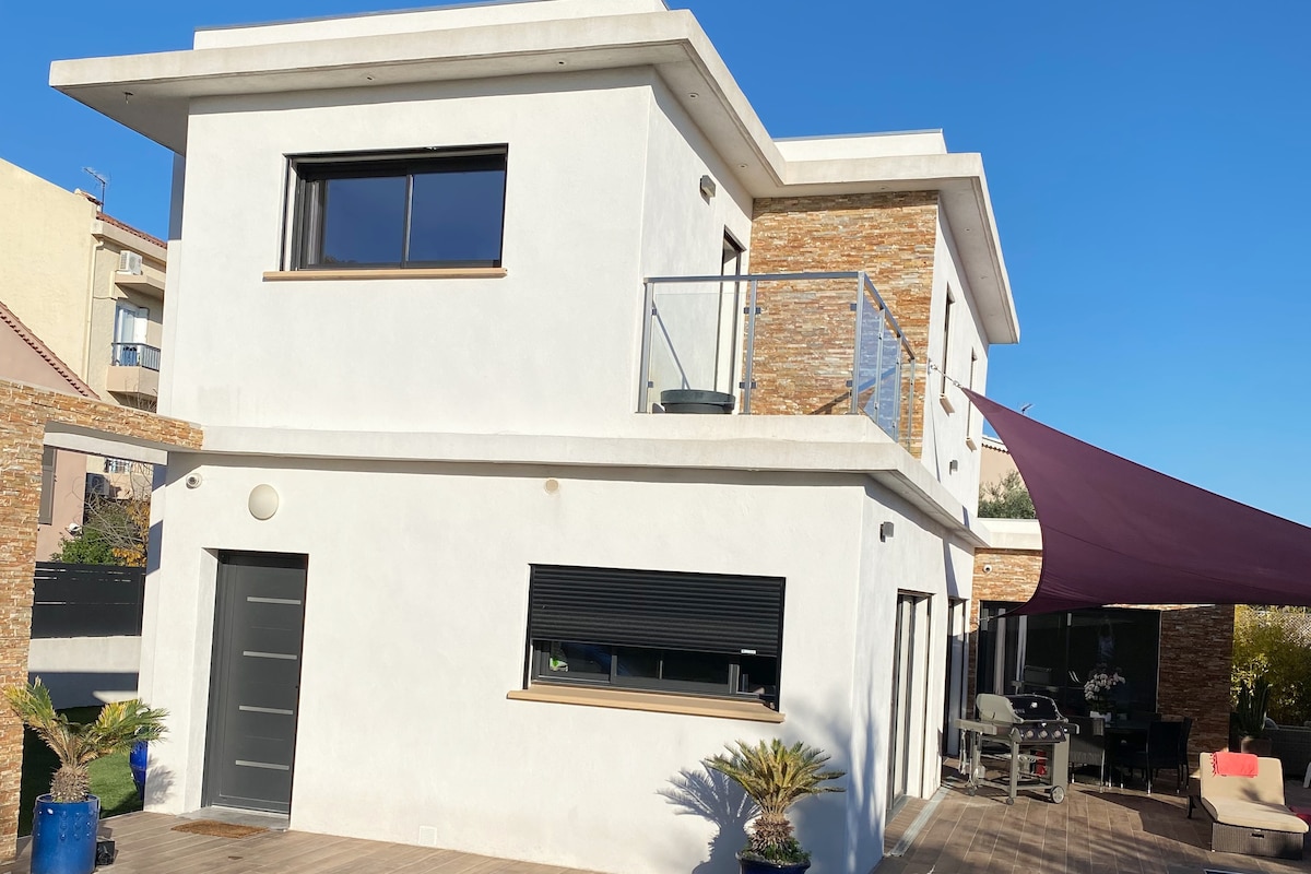The exterior of the contemporary villa showcases a combination of textured stone and smooth white walls. A balcony extends over the entrance, complemented by large windows. A shaded outdoor area with seating and a barbecue is visible, framed by palm plants and deck flooring.