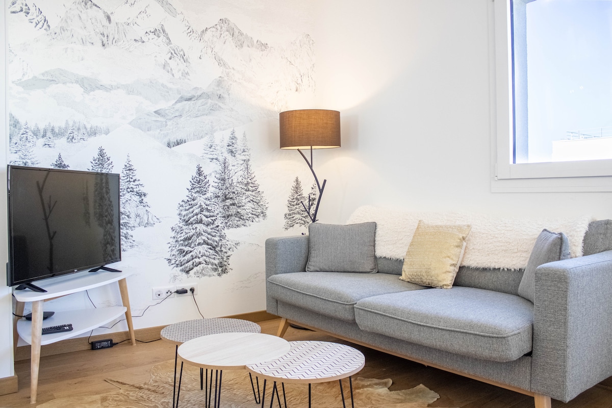 A light-filled living area features a gray sofa adorned with a soft throw pillow. Two round coffee tables sit on a wooden floor, while a TV is positioned on a white stand. A mural depicting snow-capped mountains complements the serene ambiance.