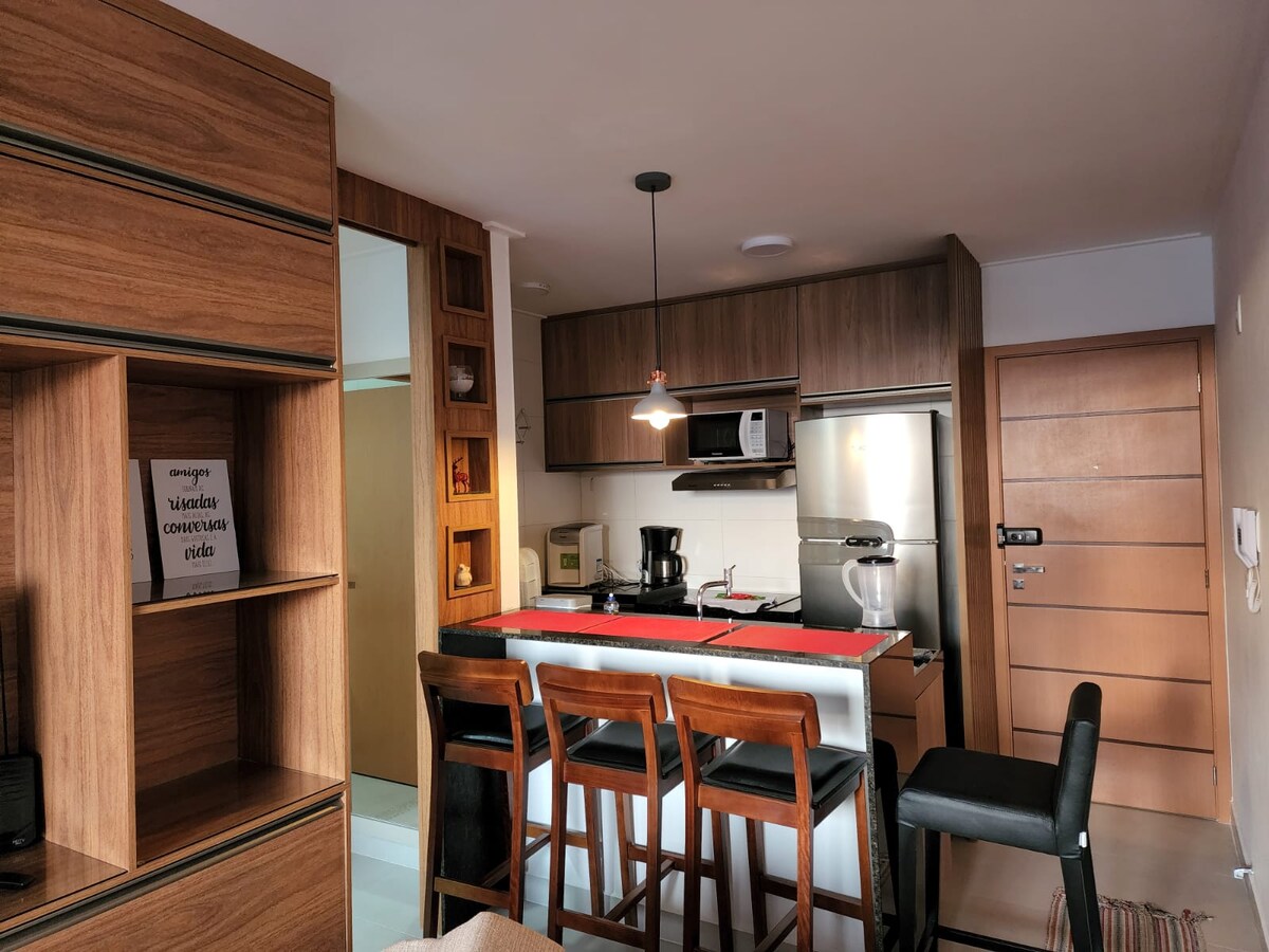 A modern kitchen is presented, featuring wooden cabinetry and a sleek countertop. A dining area with high stools is visible, and essential appliances, including a coffee maker and refrigerator, are neatly arranged. The space is well-lit, creating an inviting and functional atmosphere.