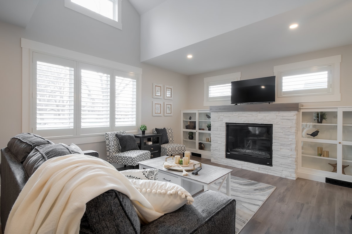 A spacious living room features comfortable seating, including a pair of patterned armchairs and a sectional sofa. Natural light enters through triple-pane windows adorned with shutters. A stone gas fireplace serves as a focal point, while shelves display decorative items. A coffee table is centrally positioned.