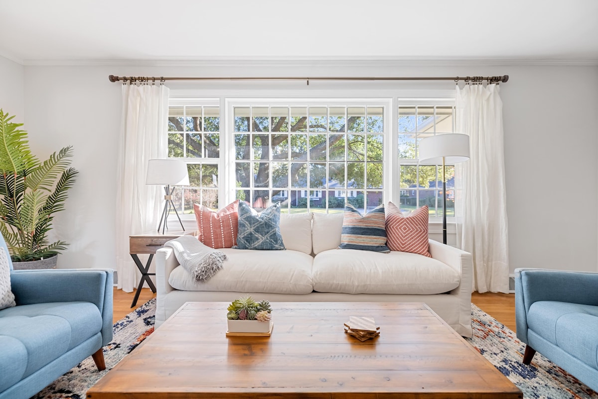A light-filled living area is furnished with a spacious white sofa adorned with patterned throw pillows in shades of blue and red. Two blue armchairs complement the decor, while a wooden coffee table with a small succulent sits at the center. Large windows overlook the yard, enhancing the inviting ambiance.