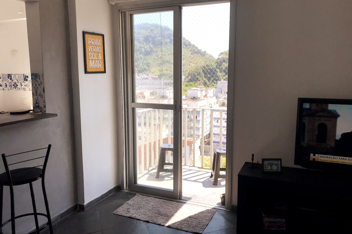 A modern living space features a sliding glass door that opens to a balcony with a scenic view of green hills. A small table and two chairs are visible on the balcony. Inside, a bar area with high chairs is positioned near the door, and a television is placed against the wall.