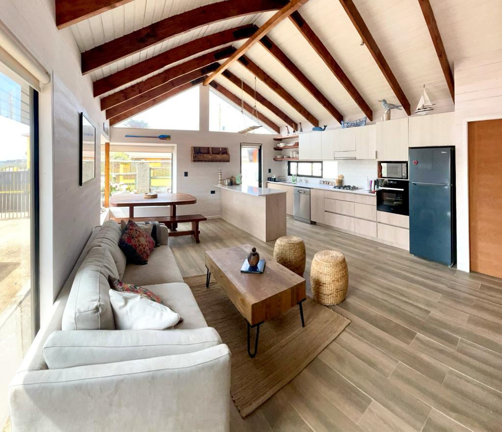 An open-concept living area features a spacious sofa with patterned cushions and a wooden coffee table centrally placed. A dining table is located nearby, and a modern kitchen with light cabinets and appliances is visible. Exposed wooden beams enhance the room's airy feel.