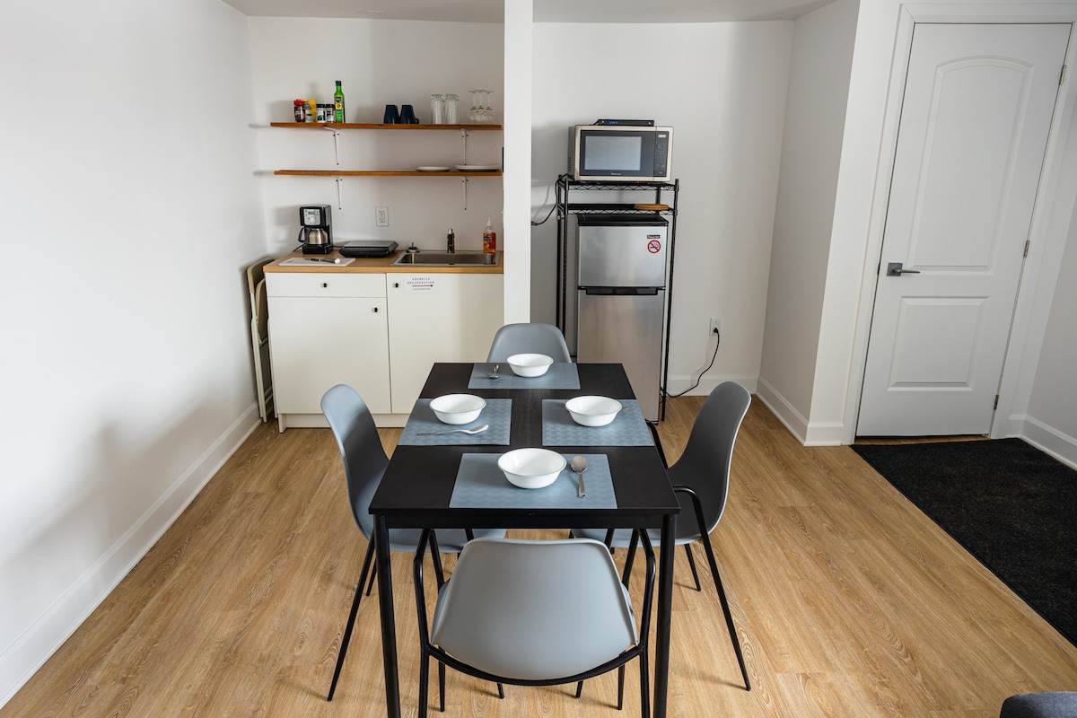 A dining area features a rectangular table set for six with plates and silverware arranged neatly. A well-equipped kitchenette is visible in the background, showcasing a microwave, refrigerator, and coffee maker on open shelving. Light wood flooring contributes to a bright environment.