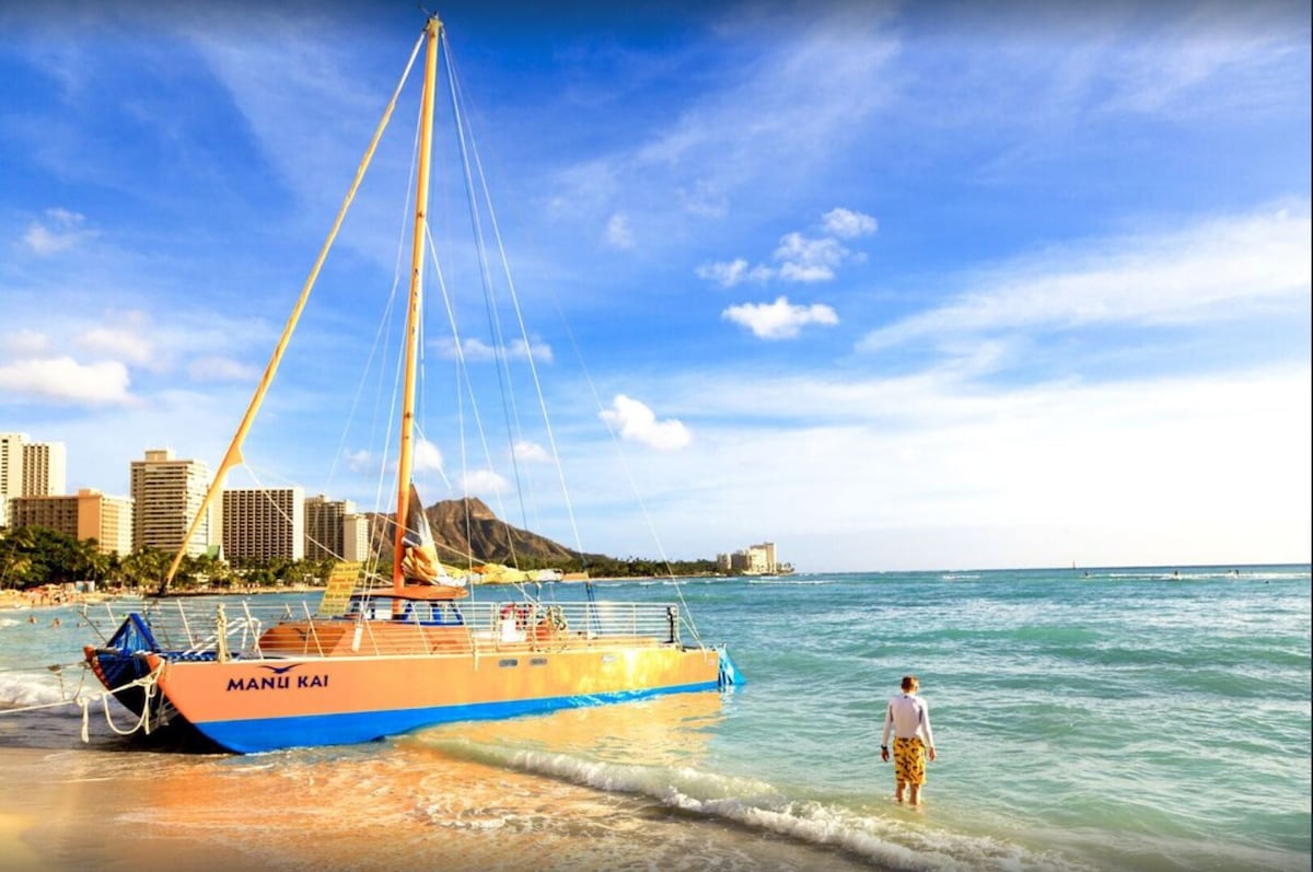 A sailboat named 'Mani Kai' is anchored just off the shore, with bright orange and blue colors contrasting against the turquoise water. Gentle waves lap at the sandy beach, while a person strolls into the calm sea. Honolulu's skyline and Diamond Head are visible in the background under a clear sky.