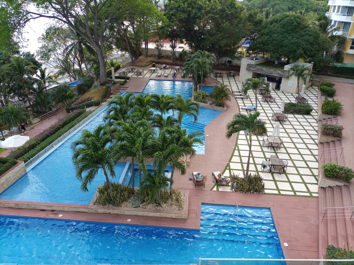 The image captures a spacious outdoor pool area surrounded by well-manicured gardens. Multiple pools are visible alongside arranged lounge chairs and tables. Lush palm trees provide shade, and clean, geometric pathways connect various sections of the space, enhancing the resort-like atmosphere.