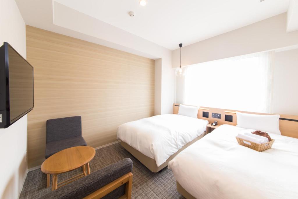 The room features two spacious beds positioned near a large window, allowing natural light to fill the space. A small wooden table and a gray chair are present, along with a flat-screen television mounted on the wall. Light-colored walls create a calming atmosphere.