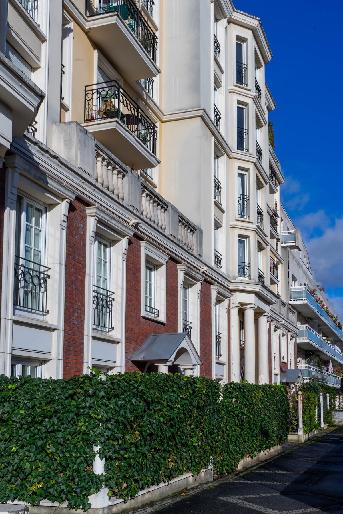 The exterior of a well-maintained building features a blend of brick and neutral-colored façades. Balconies adorned with plants line the upper floors, while manicured hedges provide privacy at ground level. The scene is set under a clear blue sky, highlighting the architectural details.