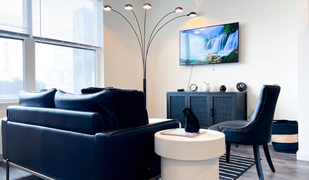 A modern living area is showcased with a black leather sofa and an accent chair beside a stylish white coffee table. Natural light filters through large windows, illuminating the space. A large TV mounted on the wall displays a serene waterfall scene.