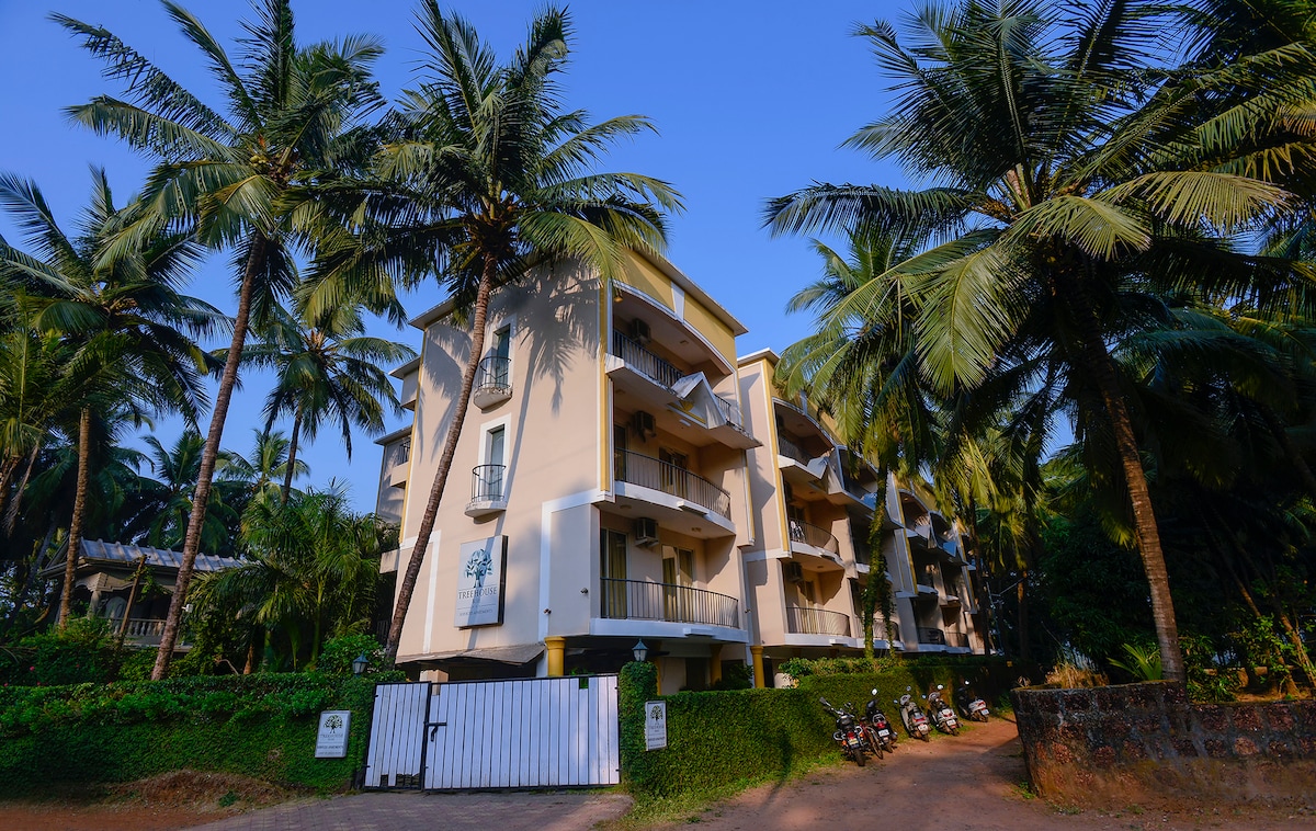 A three-story aparthotel is surrounded by lush palm trees, creating a serene atmosphere. The building features spacious balconies, with a gated entrance visible. Several parked motorcycles are present near the entrance, set against a backdrop of well-maintained greenery.