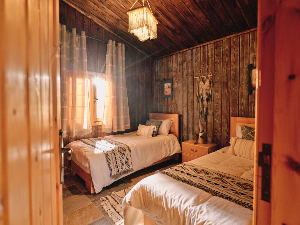 The bedroom features two twin beds with decorative bedding and ample natural light from a window dressed with sheer curtains. Wooden paneling and a rustic chandelier add warmth to the space. A small nightstand and wall decor complete the inviting atmosphere.