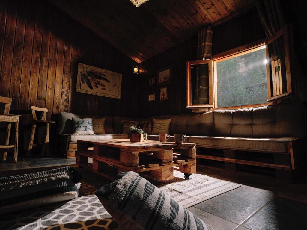 A living room area features a large, L-shaped couch made from pallets, providing ample seating. Natural light enters through a window, illuminating the wooden walls and floor. A small potted plant rests on a low coffee table, enhancing the relaxed ambiance.
