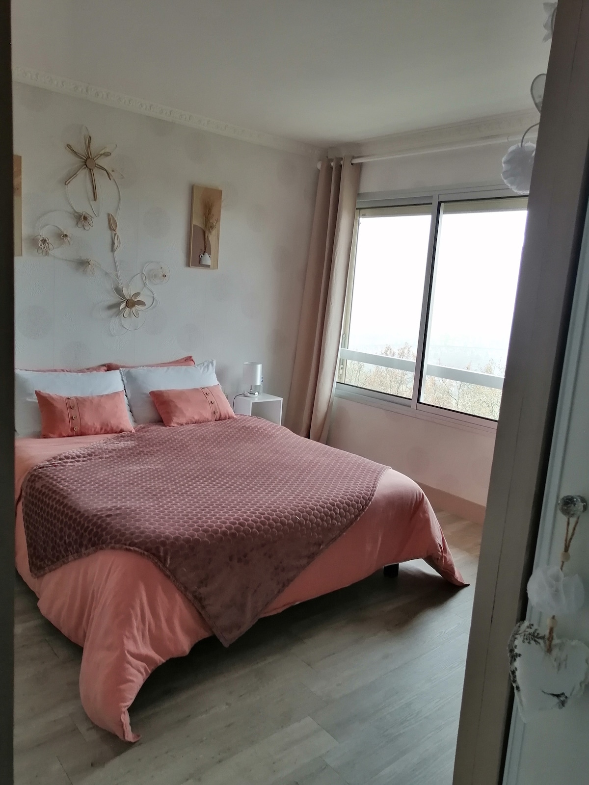 A sleeping area features a spacious bed dressed in soft coral linens, complemented by decorative cushions. Natural light floods the room through a large window, providing an expansive view of the surrounding valley. Minimalistic decor adorns the walls, enhancing the tranquil ambiance.