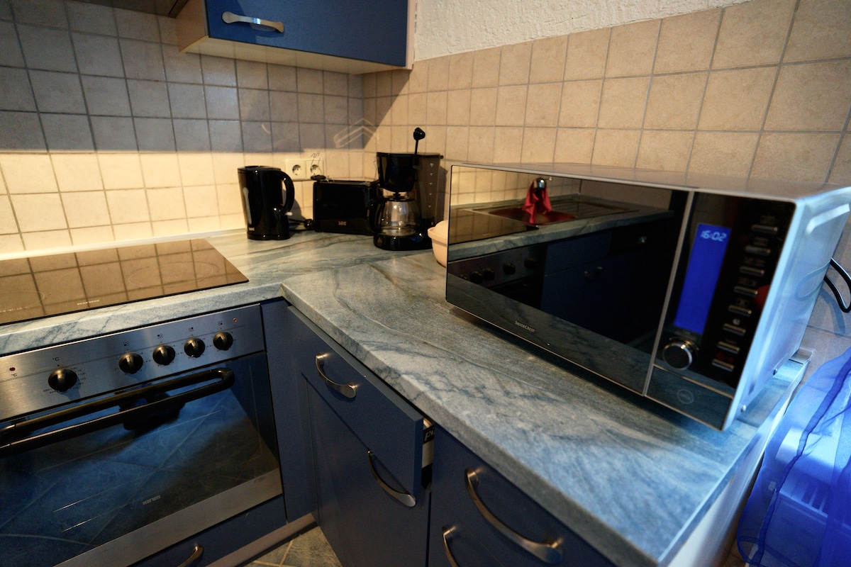 A well-equipped kitchen features modern appliances, including a microwave, coffee maker, and stovetop. Countertops are made of a blue-toned material, and beige tiled walls create a neutral backdrop. Cabinets in dark blue provide storage space, adding functionality to the design.