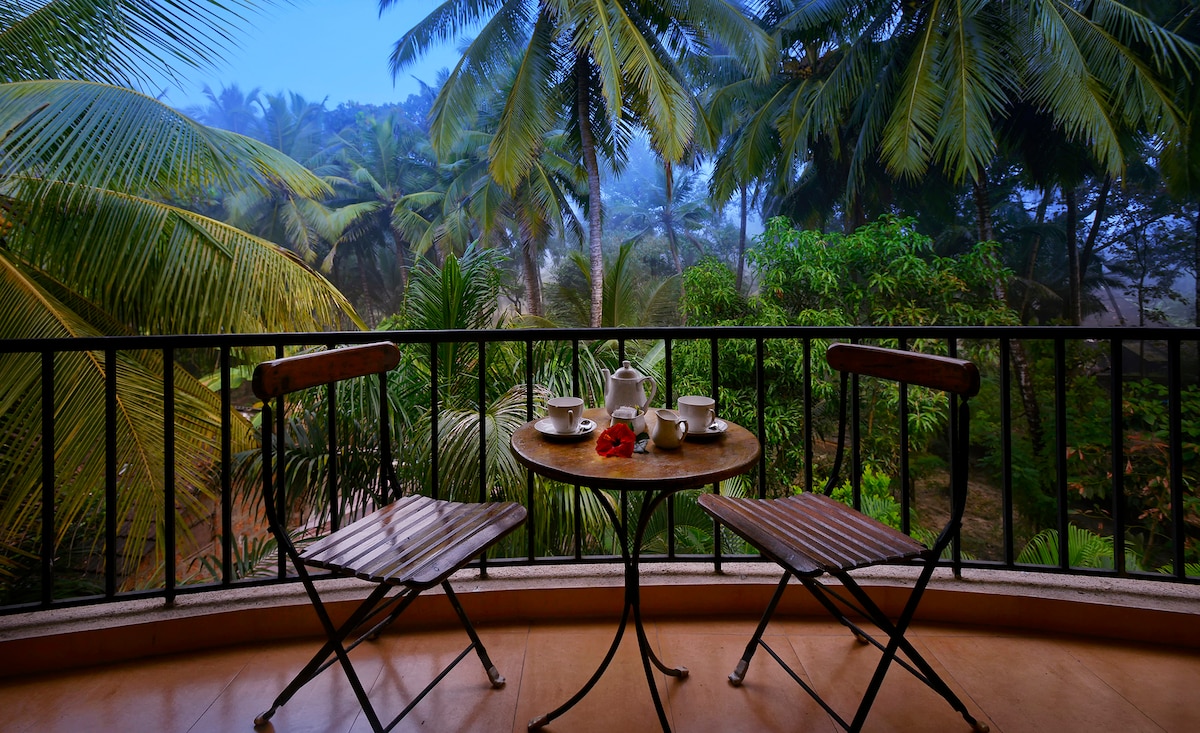 A balcony features a small round table set with two cups and a teapot, surrounded by two folding chairs. Lush green foliage and palm trees create a serene backdrop, with soft morning light filtering through the leaves.