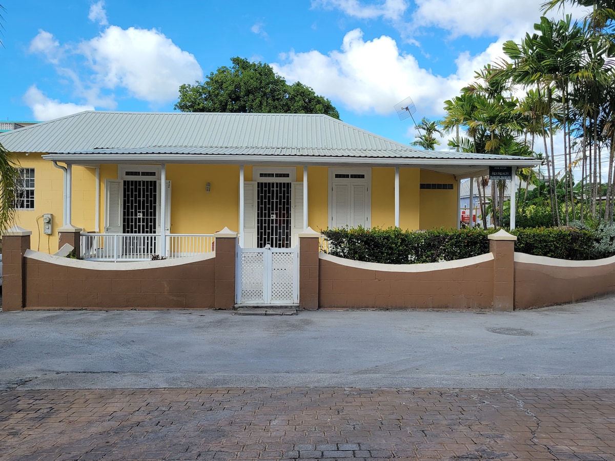 A charming beach house is showcased, featuring a yellow exterior with a green roof. The property is framed by a low brown wall and a white gate, with tropical plants visible in the front garden. A pathway leads to the entrance, set against a bright blue sky.