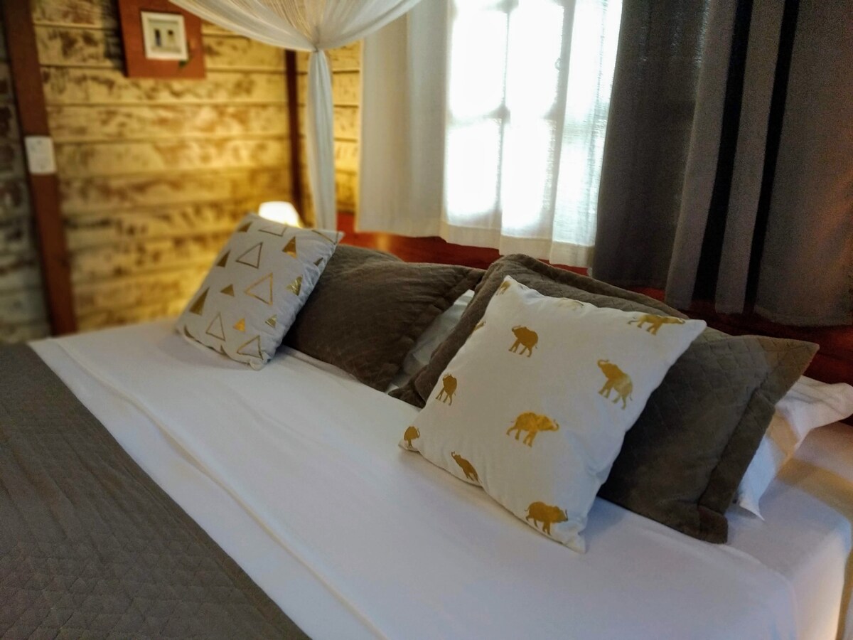 A cozy sleeping area features soft bedding layered with various decorative pillows. Light filters through sheer and darker curtains, illuminating the wooden wall paneling. A warm atmosphere is created by the mix of textures and a serene color palette.