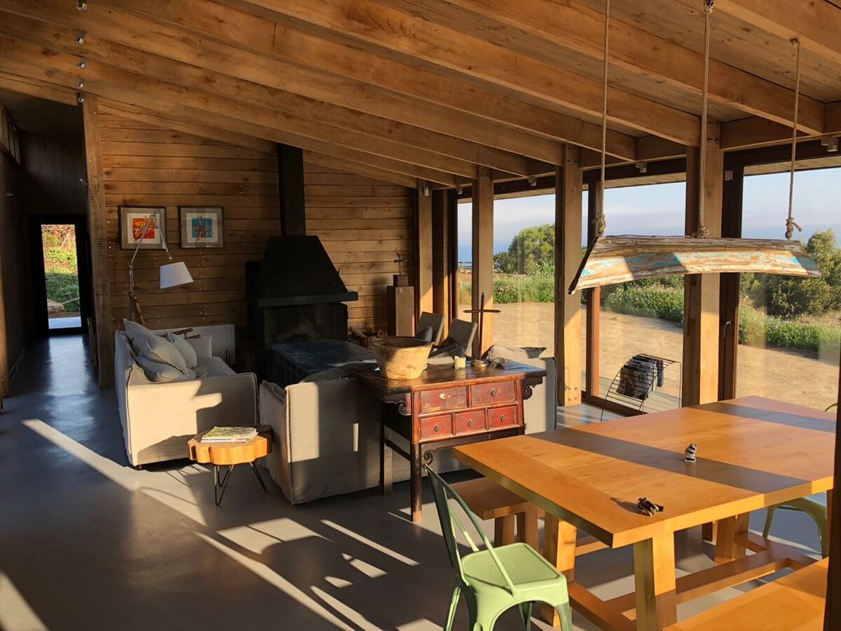 An open-concept living area features high wooden ceilings and large windows offering views of the landscape. A cozy seating arrangement with light-colored couches is positioned near a fireplace, complemented by a wooden dining table surrounded by green chairs.