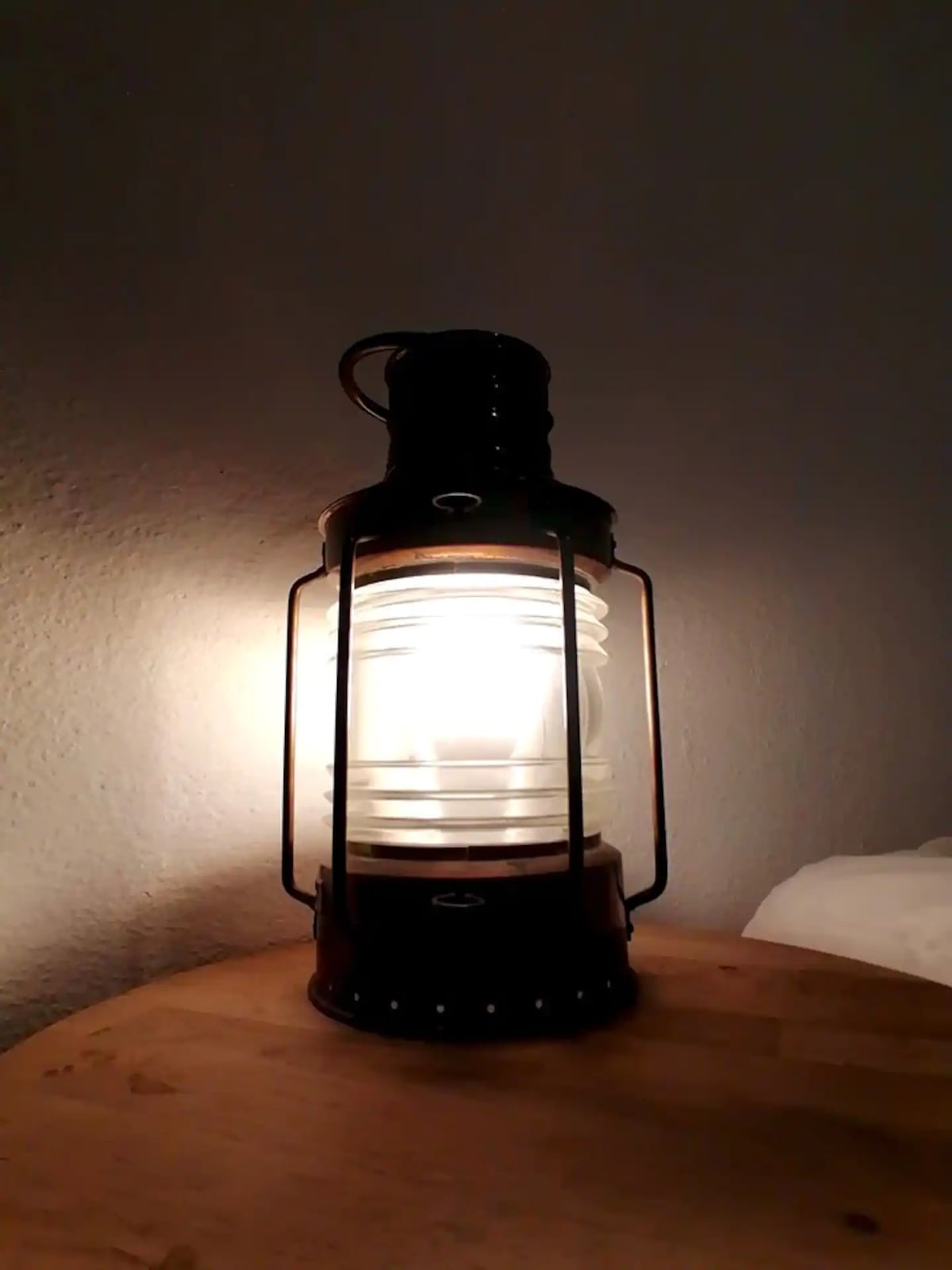 A vintage lantern emits a warm light, casting a soft glow against the textured wall. The lantern is positioned on a wooden surface, complemented by nearby bedding that offers an inviting touch to the space.