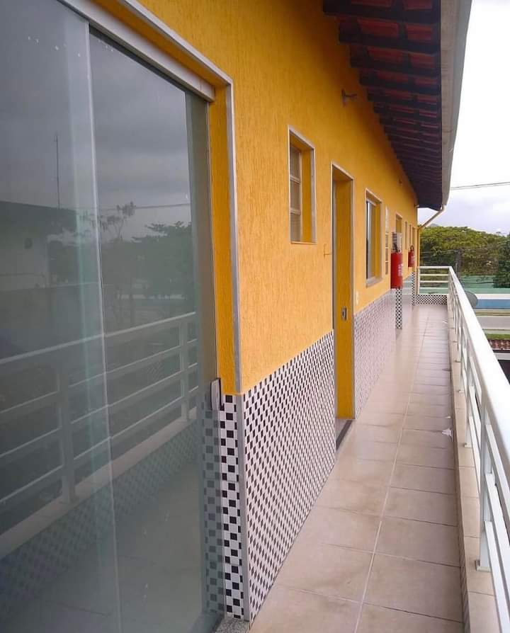 A balcony lined with a pale tile floor features a vibrant yellow exterior wall. The space includes several windows, with sliding glass doors leading to individual rooms. Checkered tile accents near the base provide a contrasting detail along the lower wall.