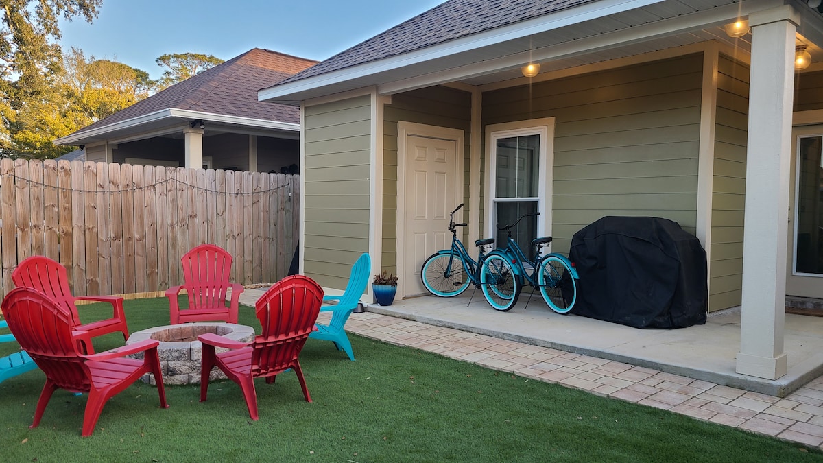 An outdoor seating area features a circular arrangement of red Adirondack chairs around a stone firepit, set on a grassy surface. Two beach cruiser bikes are positioned beside a gas grill, with a fenced yard and exterior door visible in the background.