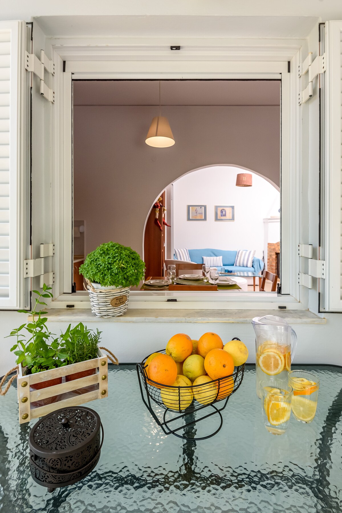 A kitchen window opens to a cozy living area, where a blue couch and framed artwork can be seen. A table is set with a bowl of oranges and lemons, along with a pitcher of lemonade and a decorative container of herbs, creating a fresh ambiance.