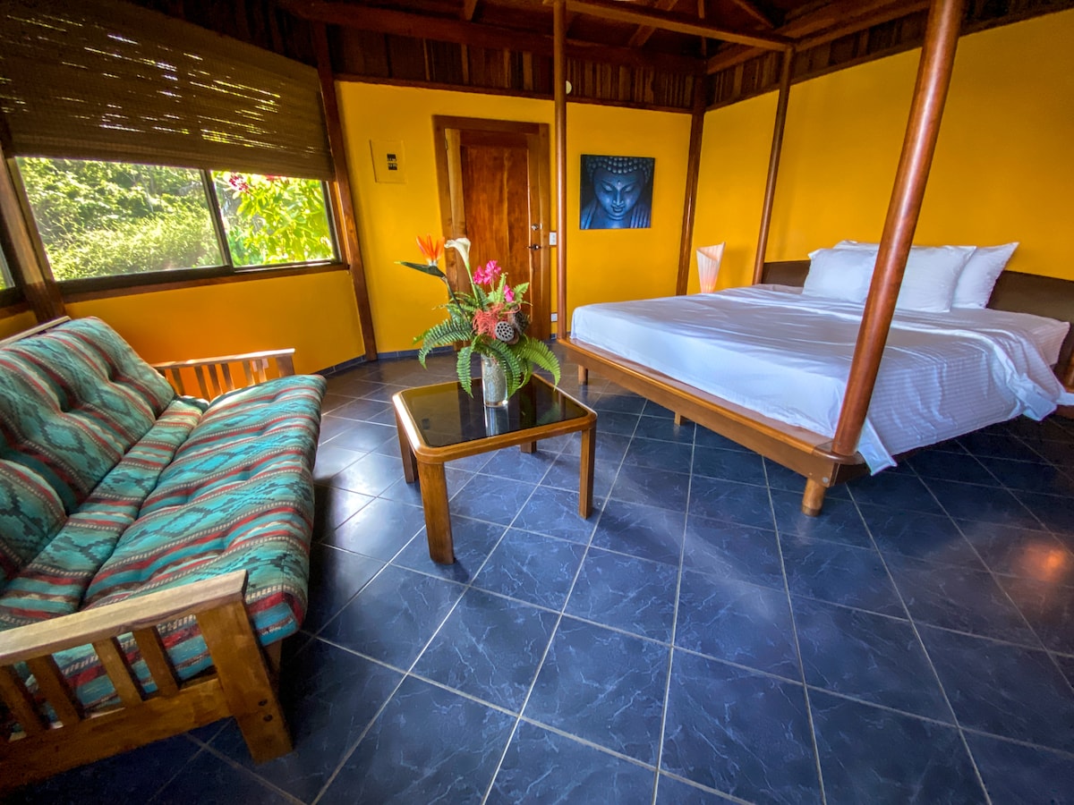 A spacious room features a king bed with crisp linens, complemented by a cozy seating area with a patterned sofa and a coffee table. Natural light filters in through large windows, highlighting the vibrant yellow walls and lush outdoor view.