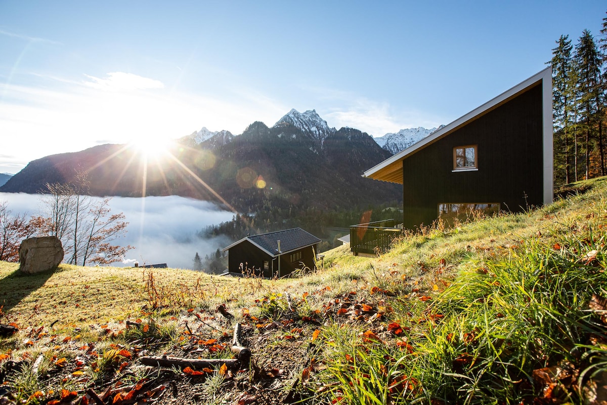 The cabin is positioned amidst gently sloping grass, while the sun rises behind majestic mountains, illuminating a mist-covered valley. Nearby trees frame the scene, with a second cabin visible in the background, creating a serene atmosphere.