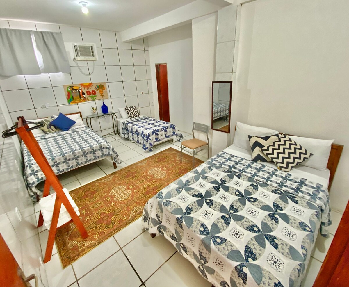 A spacious guest room features two beds adorned with patterned bedding, providing comfortable sleeping arrangements for guests. A traditional rug complements the tiled floor, while a wall mirror enhances the room's openness. An air conditioning unit and a small desk are also visible.