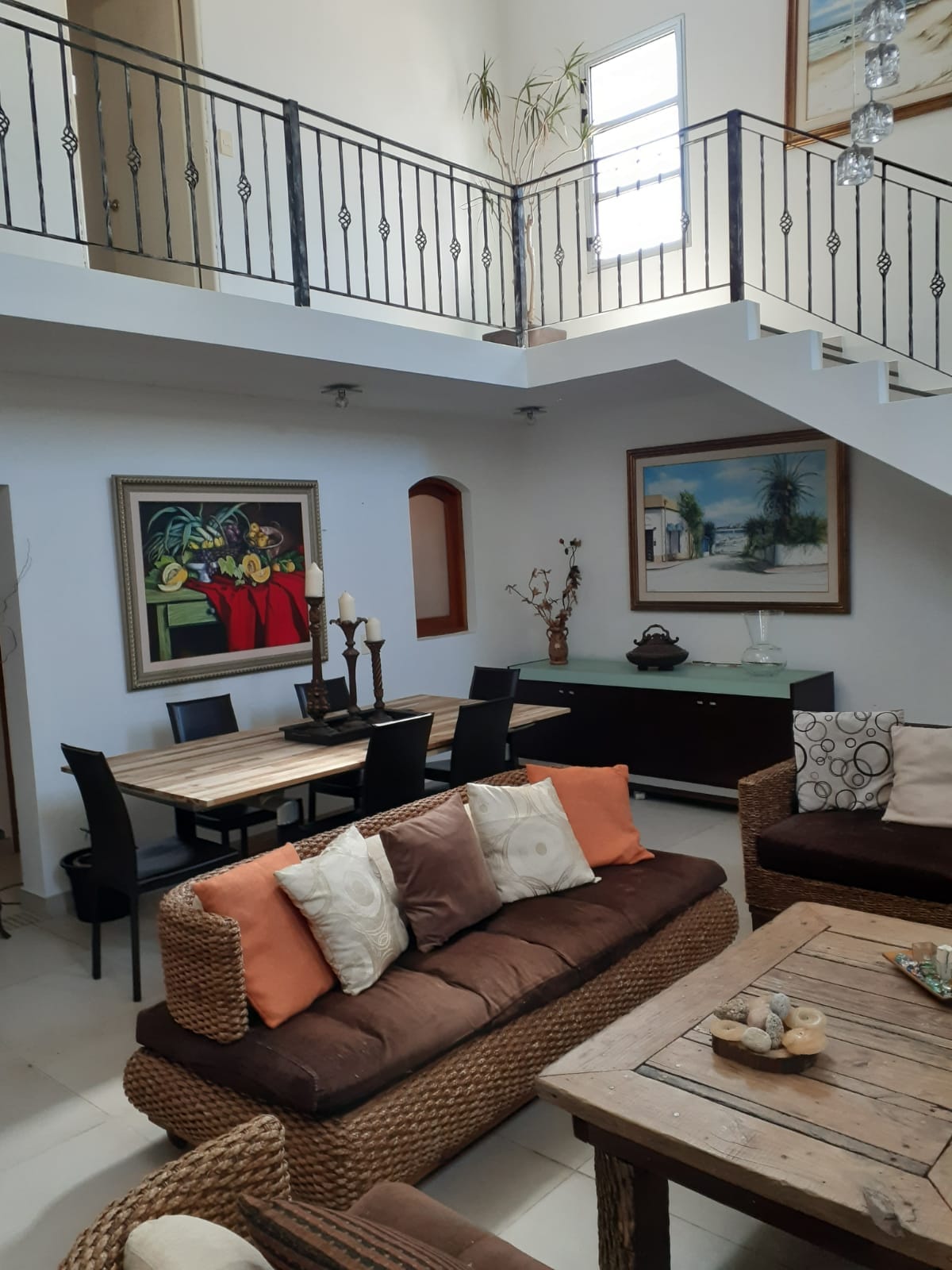 An airy living space is captured, featuring a mix of seating options with a woven sofa adorned with orange and cream cushions. A wooden dining table with black chairs sits in proximity, while artwork enhances the walls. Natural light illuminates the area from high windows.