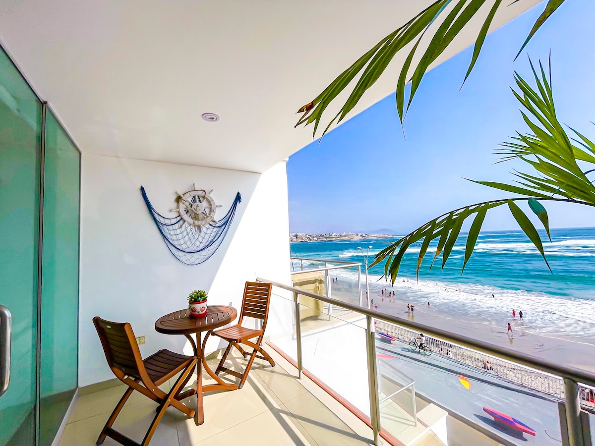 A balcony view featuring a round table and two wooden chairs, with a decorative nautical piece on the wall. Waves can be seen crashing onto the shore below, while people walk along the beach. Green palm leaves frame the scene, enhancing the beachfront perspective.