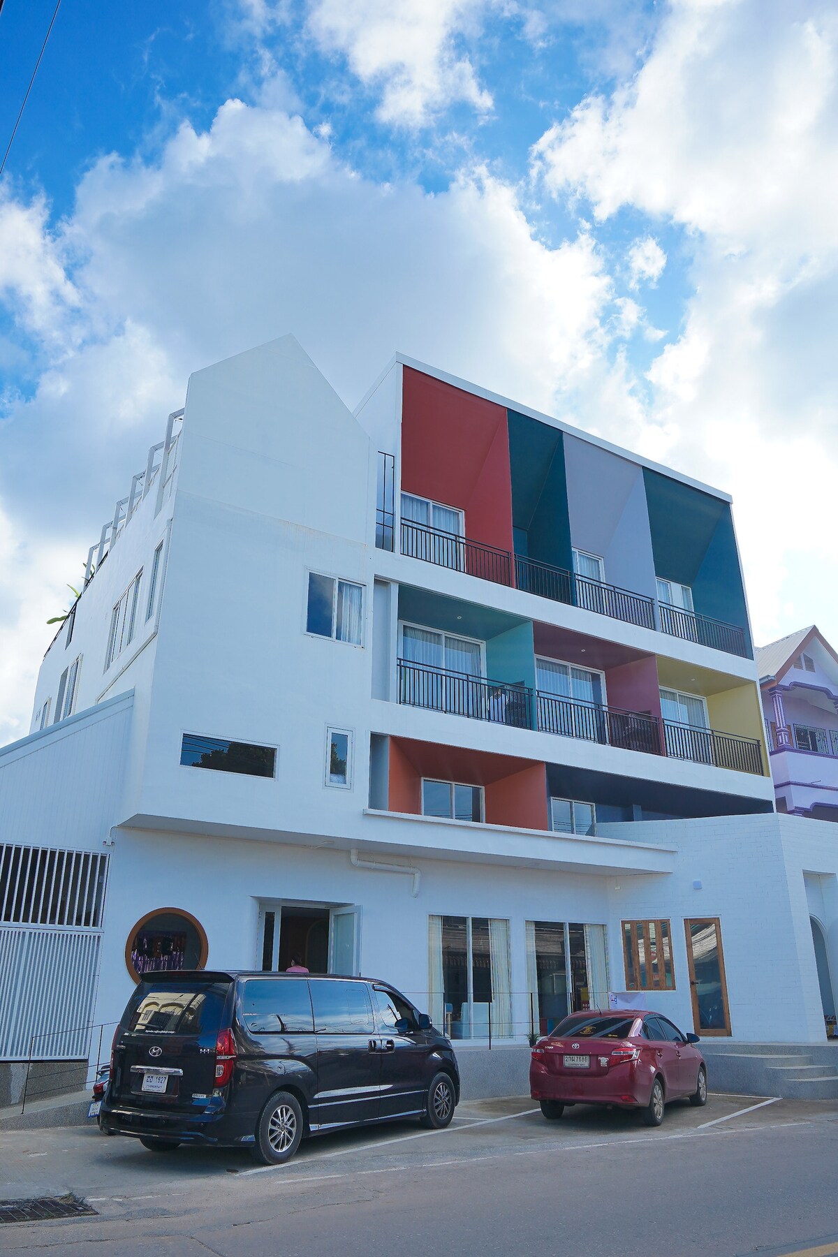 A modern, three-story building features a clean, white exterior accented by vibrant colors in geometric patterns. Balconies with railings are visible on each floor. Two parked cars are located in front of the entrance, with an expansive sky filled with clouds overhead.