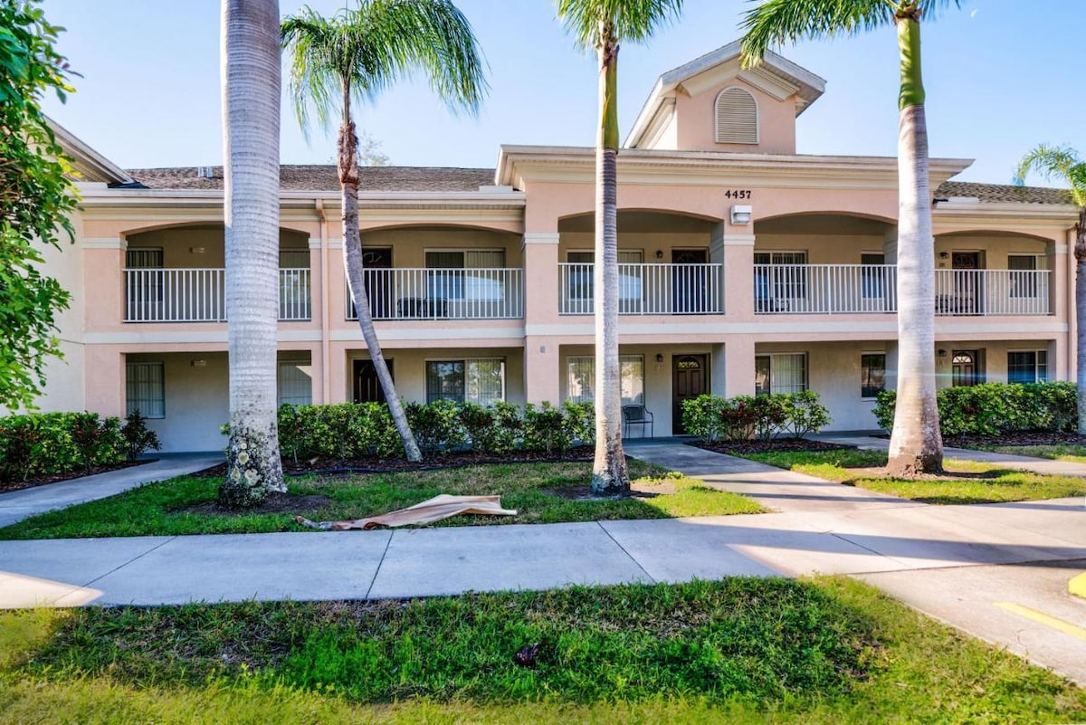 A well-maintained two-story building is framed by palm trees and lush greenery. The entrance is highlighted by a covered balcony with white railings. A manicured walkway leads to the doorway, creating an inviting access point into the community.