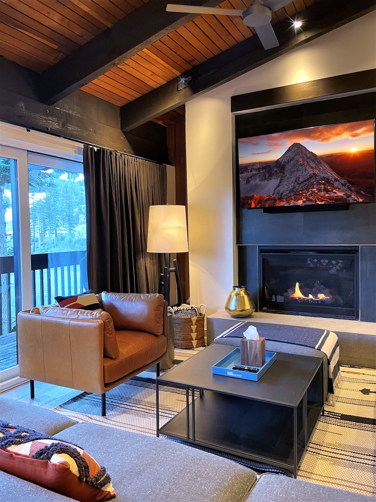 The living room showcases a warm and inviting atmosphere, featuring a cozy seating area with a leather armchair and modern sofa. A gas fireplace is complemented by a flat-screen TV, while sliding glass doors provide access to a spacious balcony with views of the surrounding nature.