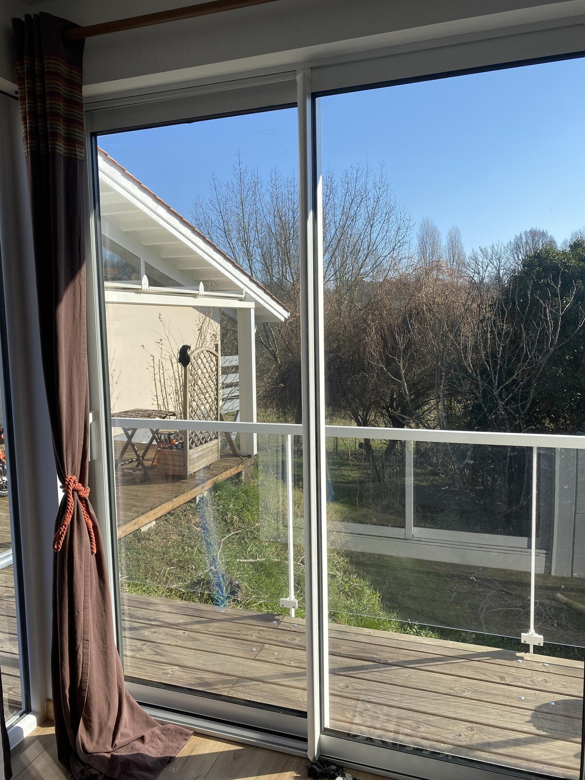 A large sliding glass door offers a view of a balcony and a natural landscape. Sunlight enters the space, illuminating the area with a bright and airy ambiance. The balcony features a railing and is surrounded by trees and greenery.