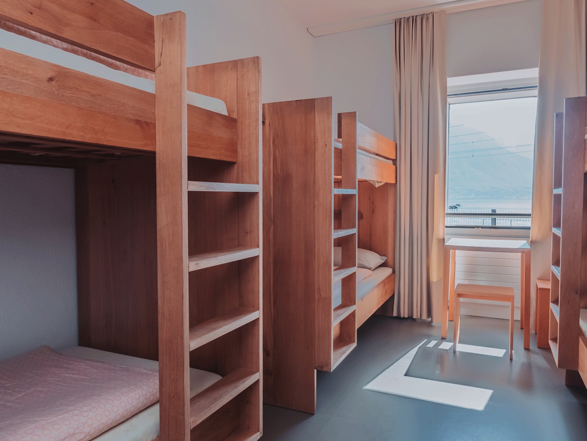 A spacious room features two sets of wooden bunk beds, providing ample sleeping areas. A simple wooden chair is positioned next to a window, adorned with light curtains that allow natural light to filter in, highlighting the clean, minimalist design.