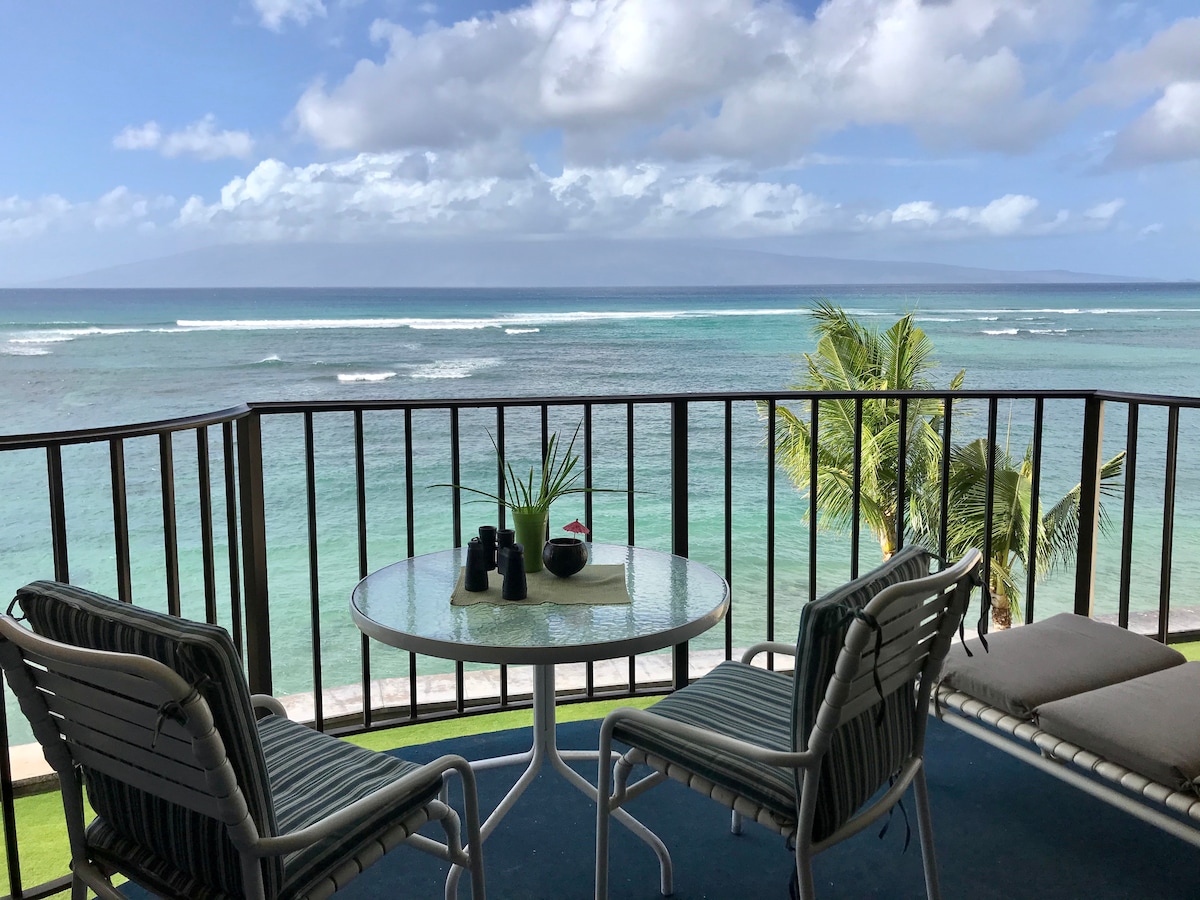 A private lanai features a round table set with decorative items, surrounded by two striped chairs. The serene ocean view showcases gentle waves and a distant island under a sky with scattered clouds, complemented by a nearby palm tree swaying in the breeze.