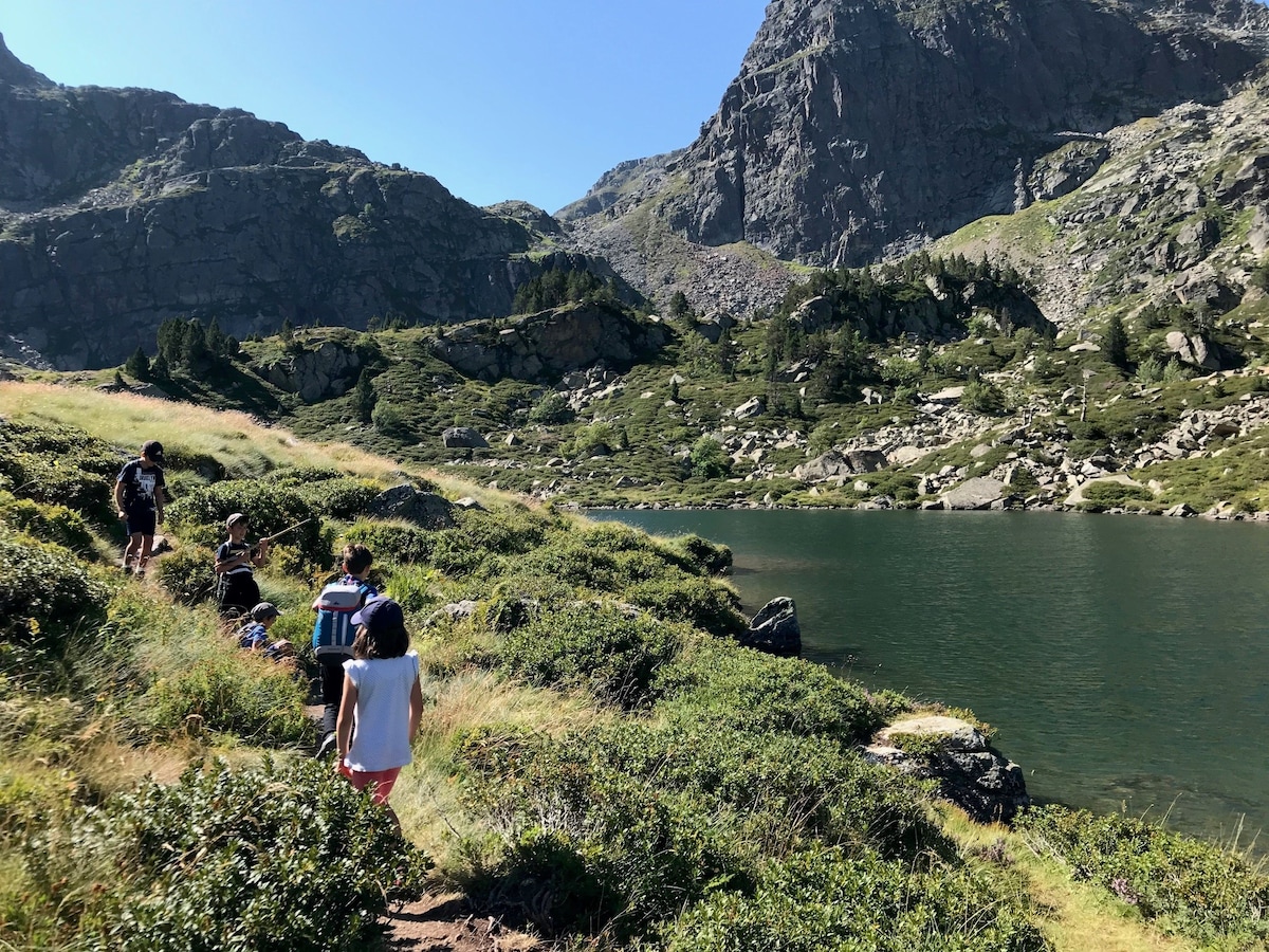 A serene landscape features a group of individuals walking along a grassy bank beside a calm lake. Rocky mountains rise in the background, framed by vibrant greenery and patches of sunlight, showcasing the natural beauty of the surrounding area.