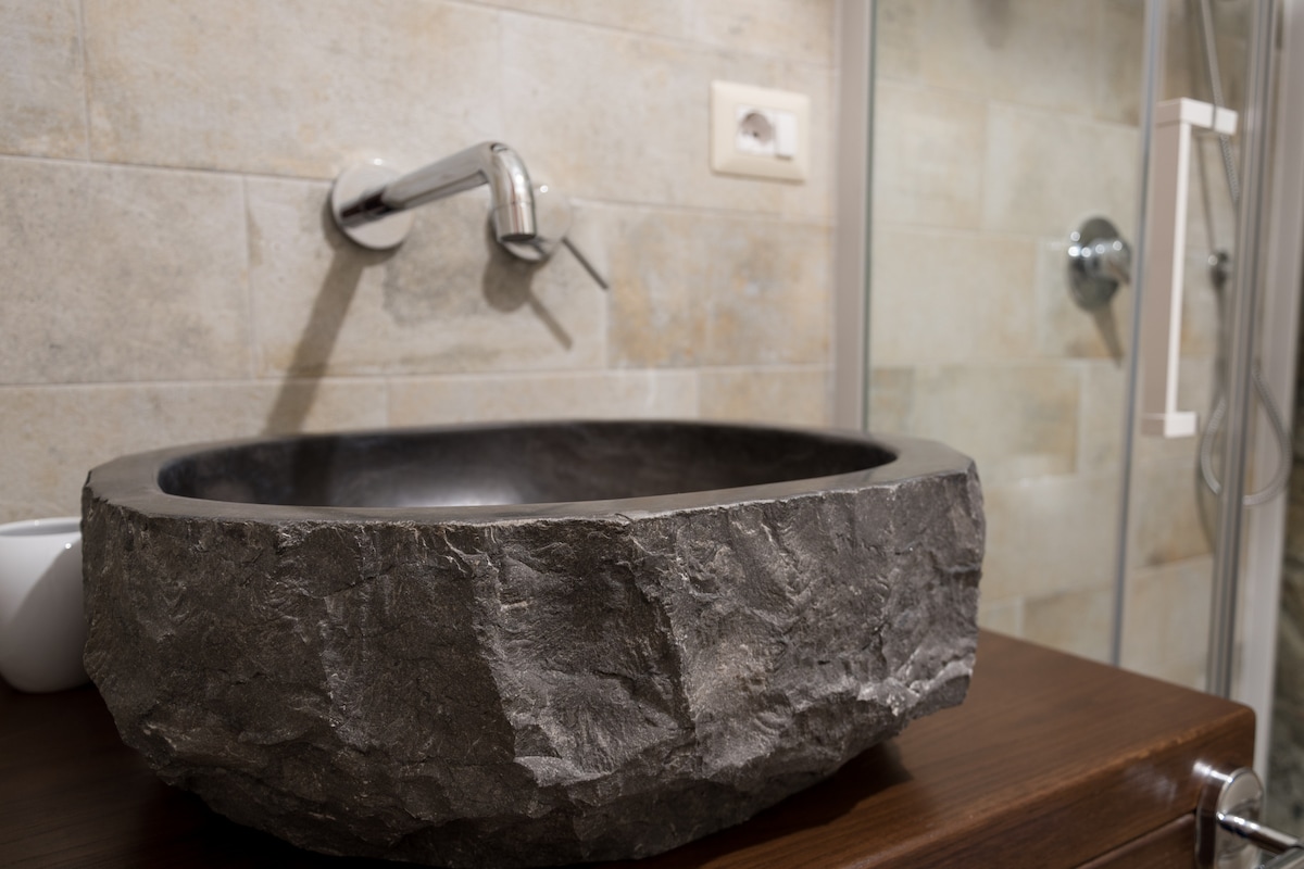 A unique stone sink is displayed on a wooden vanity, showcasing its natural texture. The light grey tiles adorn the walls, complementing the sink. A simple faucet extends from the wall, and a glass shower is partially visible in the background.