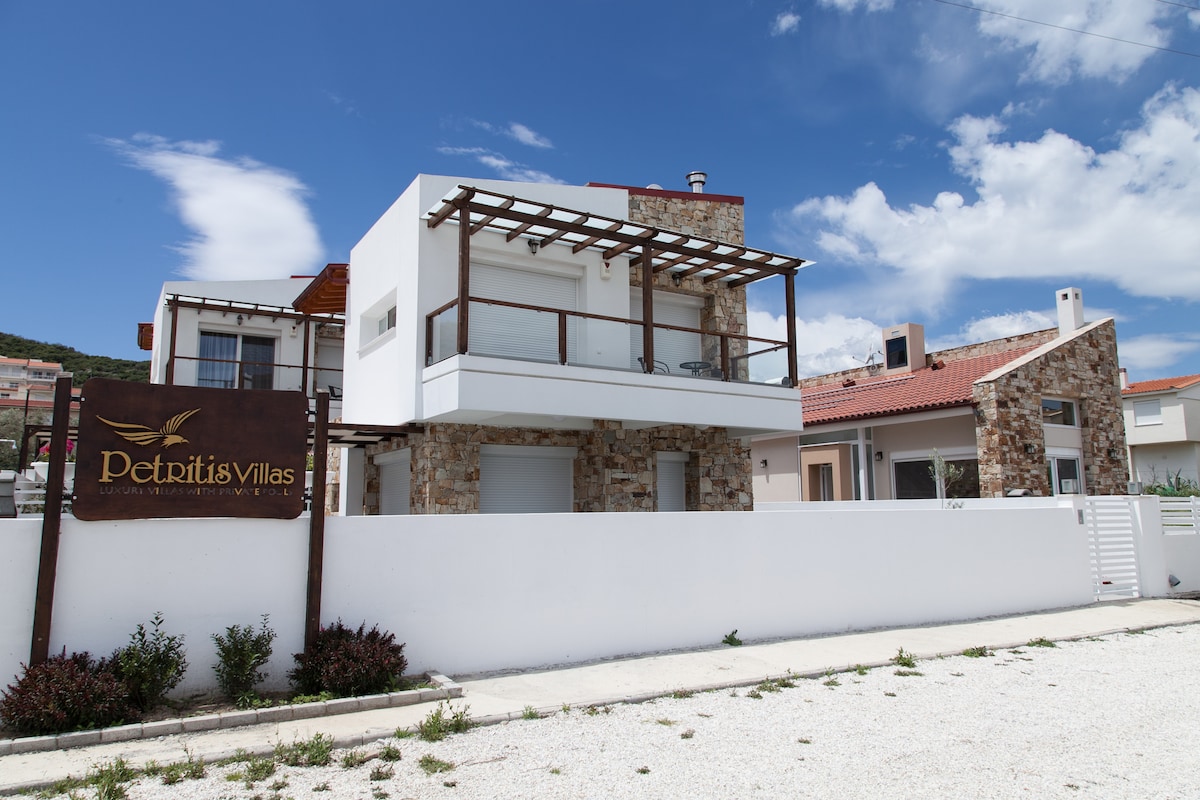 A modern villa with a stone facade and wooden accents is displayed. A covered balcony is visible above the entrance, complemented by a well-maintained garden area. The white exterior walls contrast with the blue sky, enhancing the villa's contemporary architecture.
