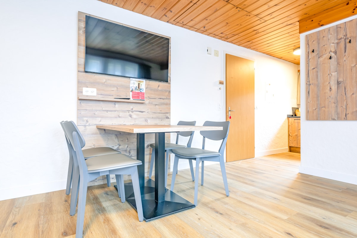 A dining area is presented with a rectangular table surrounded by four gray chairs. A wall-mounted flat-screen TV is visible above a rustic wooden panel. Light hardwood flooring enhances the bright and open feel of the space.