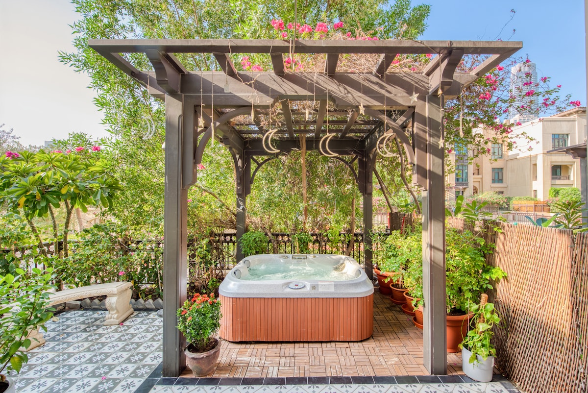 A private garden features a wooden pergola, under which a personal jacuzzi is situated. Surrounding greenery, including flowering plants, enhances the outdoor space. A decorative tiled surface provides a contrasting foundation, complementing the tranquil ambiance.