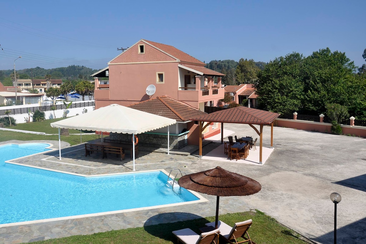 A communal outdoor swimming pool is featured prominently, surrounded by sun loungers and umbrellas. A shaded area with a thatched roof provides seating next to the pool. The surrounding landscaped area includes green trees and features a multi-story building in the background.