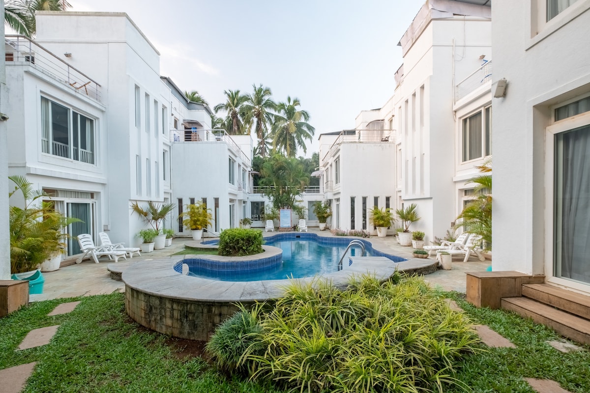 A serene courtyard features a shared swimming pool surrounded by white buildings. Palm trees and lush greenery create a peaceful ambiance. Lounge chairs are positioned around the pool, inviting relaxation amidst a tranquil setting.