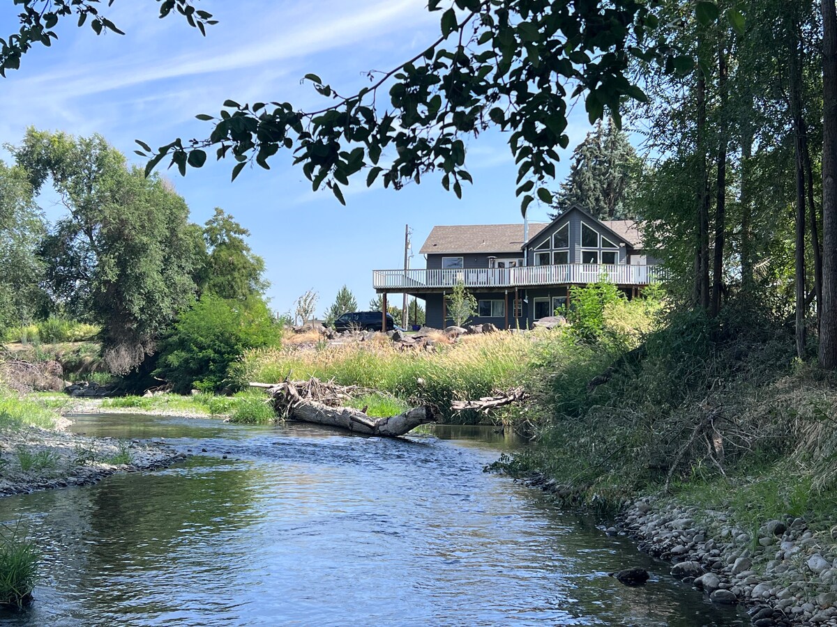 The two-story house is set on a slight elevation above the river, presenting large windows and a spacious deck. Surrounding greenery includes trees and grass, with a gentle river flowing in the foreground, offering a serene natural landscape.