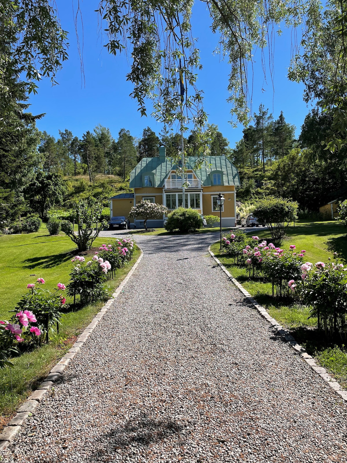 A gravel path leads towards a charming yellow house with a glass-roofed structure, surrounded by lush greenery. Flowerbeds with blooming peonies line the route, enhancing the natural beauty of the landscaped garden.