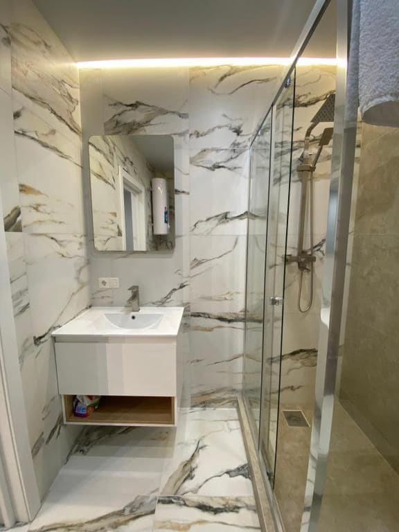 A modern bathroom features elegant marble-patterned walls and a glass-enclosed shower. A sleek white vanity with an open shelf for storage complements the design. Natural light enters the space through a window, enhancing the airy feel.
