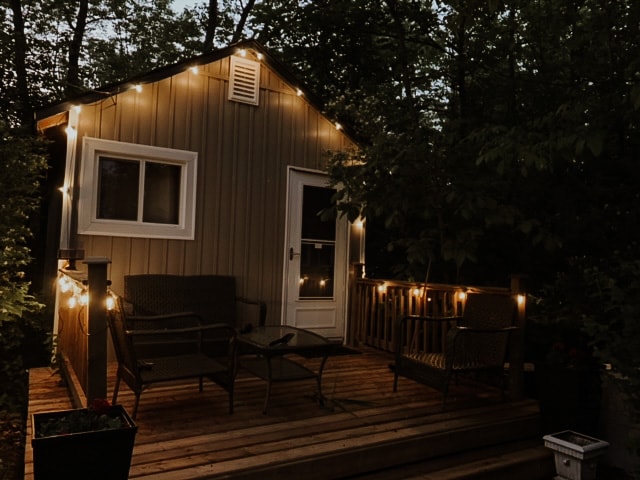 A cozy outdoor seating area is illuminated by warm string lights. Two comfortable chairs and a glass table are arranged on a wooden deck, surrounded by lush greenery. A door leads into the cottage, providing an inviting entrance.
